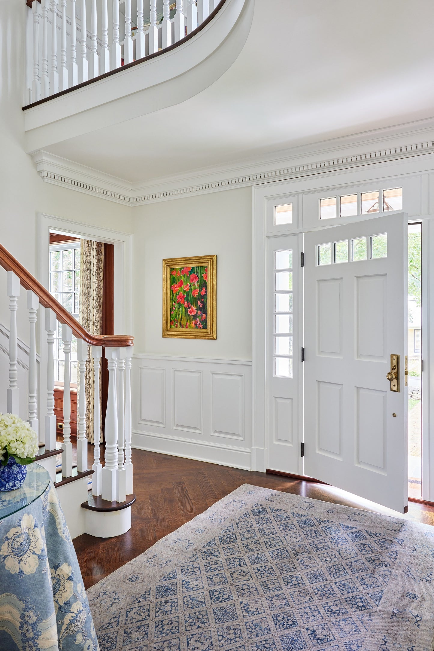 Bright traditional entryway with white panel walls, wooden staircase, blue patterned rug, gold-framed painting near the front door.