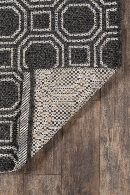 Dark gray rug with white geometric octagon and square pattern, folded corner reveals beige underside, wood floor backdrop.