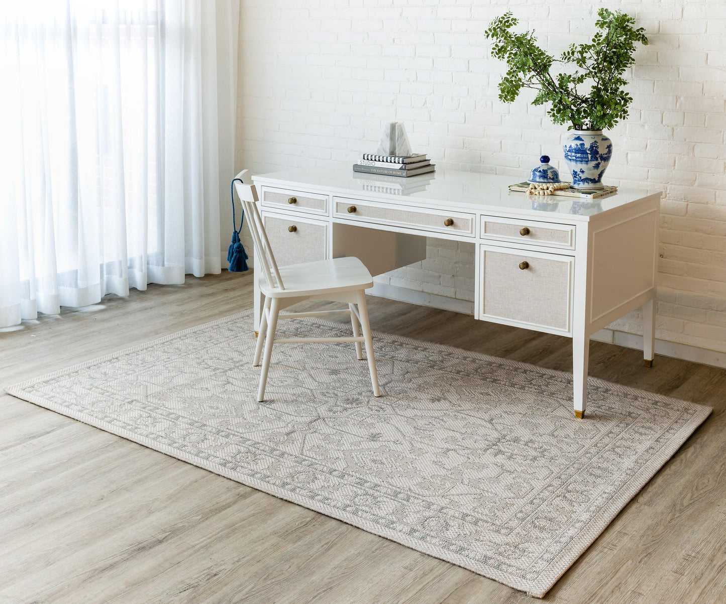 White wooden desk with drawers, matching chair, light gray patterned rug, brick wall, blue-and-white vase with greenery.