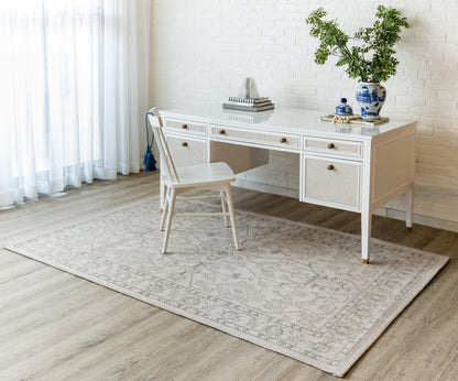 White wooden desk with drawers, matching chair, light gray patterned rug, brick wall, blue-and-white vase with greenery.