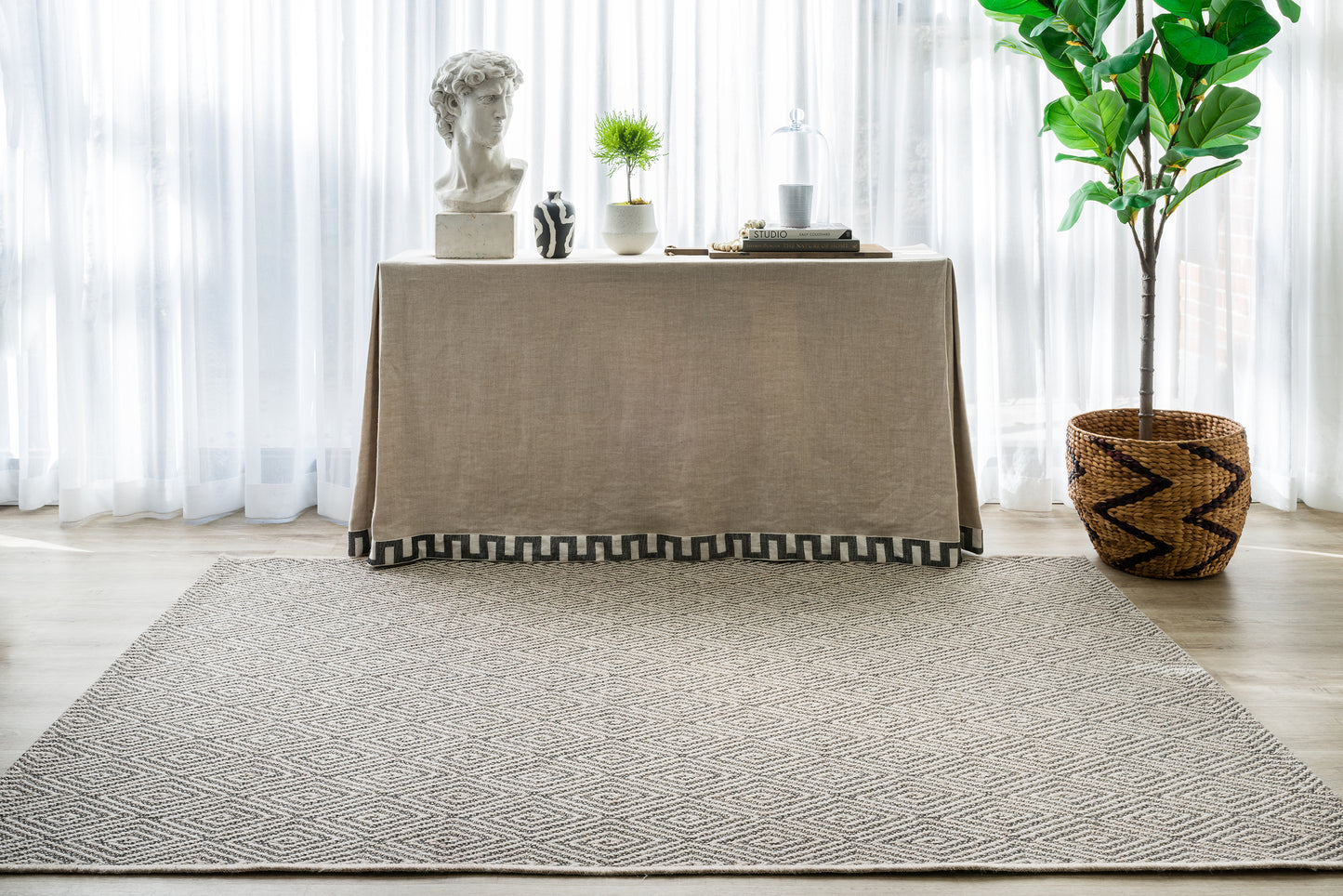 Beige linen-draped console with bust, plants and books; large gray textured rug and woven basket plant indoors.