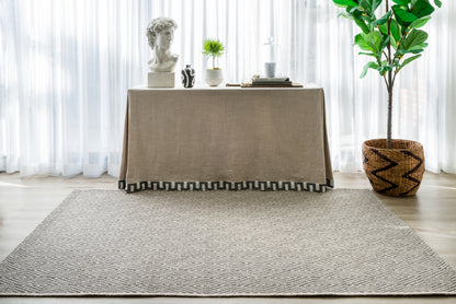 Beige linen-draped console with bust, plants and books; large gray textured rug and woven basket plant indoors.