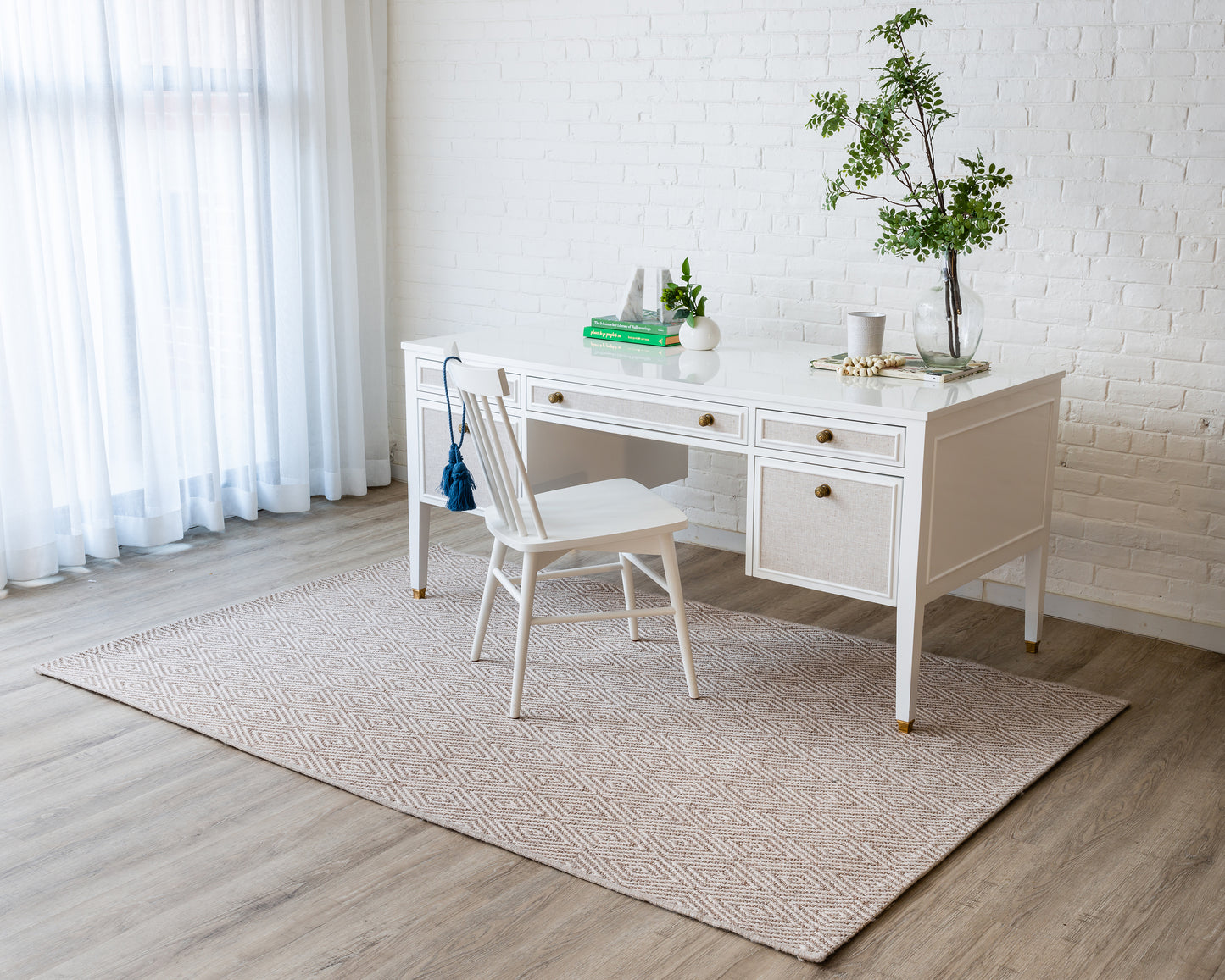 White wooden desk with woven drawer fronts and brass knobs, matching white chair, beige geometric rug in bright home office.