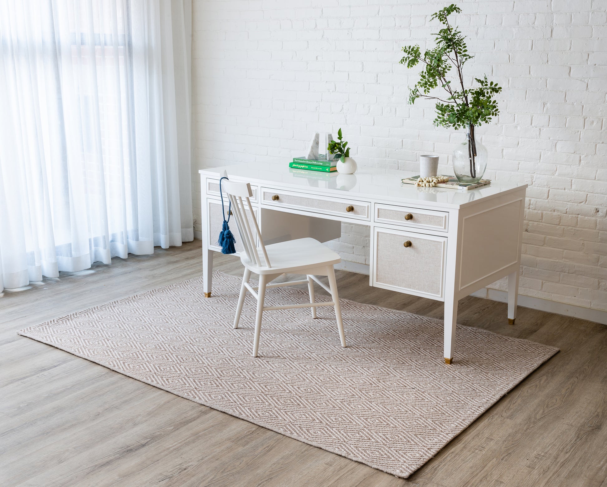 White wooden desk with woven drawer fronts and brass knobs, matching white chair, beige geometric rug in bright home office.