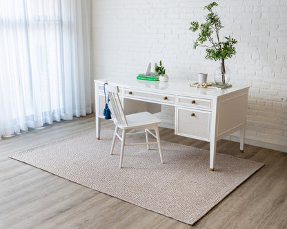White wooden desk with woven drawer fronts and brass knobs, matching white chair, beige geometric rug in bright home office.