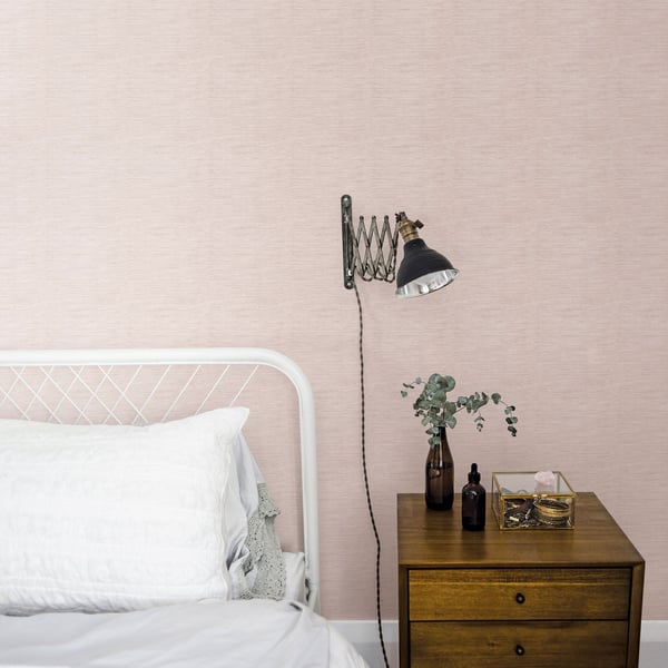 Blush pink textured wallpaper, white metal bed frame, industrial wall lamp, wooden two-drawer nightstand with vase and bottles.