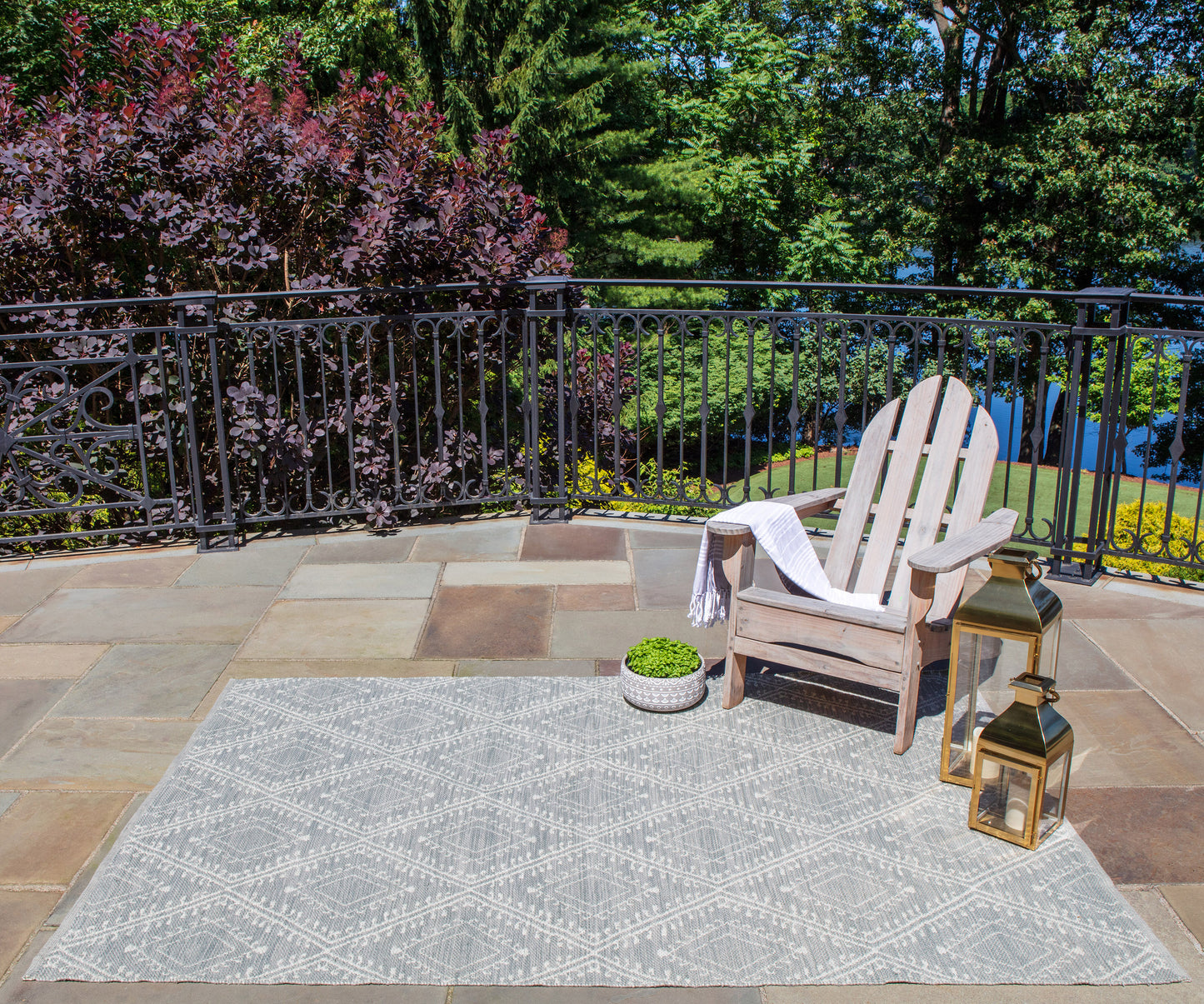 Outdoor patio: gray geometric rug, wooden Adirondack chair with towel, potted plant, and gold lanterns by black railing.