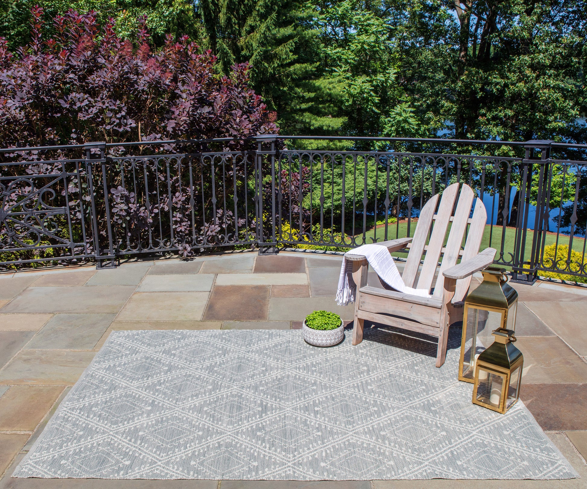 Outdoor patio: gray geometric rug, wooden Adirondack chair with towel, potted plant, and gold lanterns by black railing.