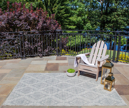 Outdoor patio: gray geometric rug, wooden Adirondack chair with towel, potted plant, and gold lanterns by black railing.
