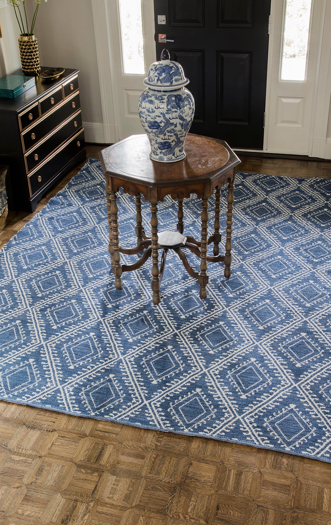 Blue geometric rug, 5' x 8', on the floor in a hallway; a blue & white porcelain vase sits on a round carved-wood side table nearby.