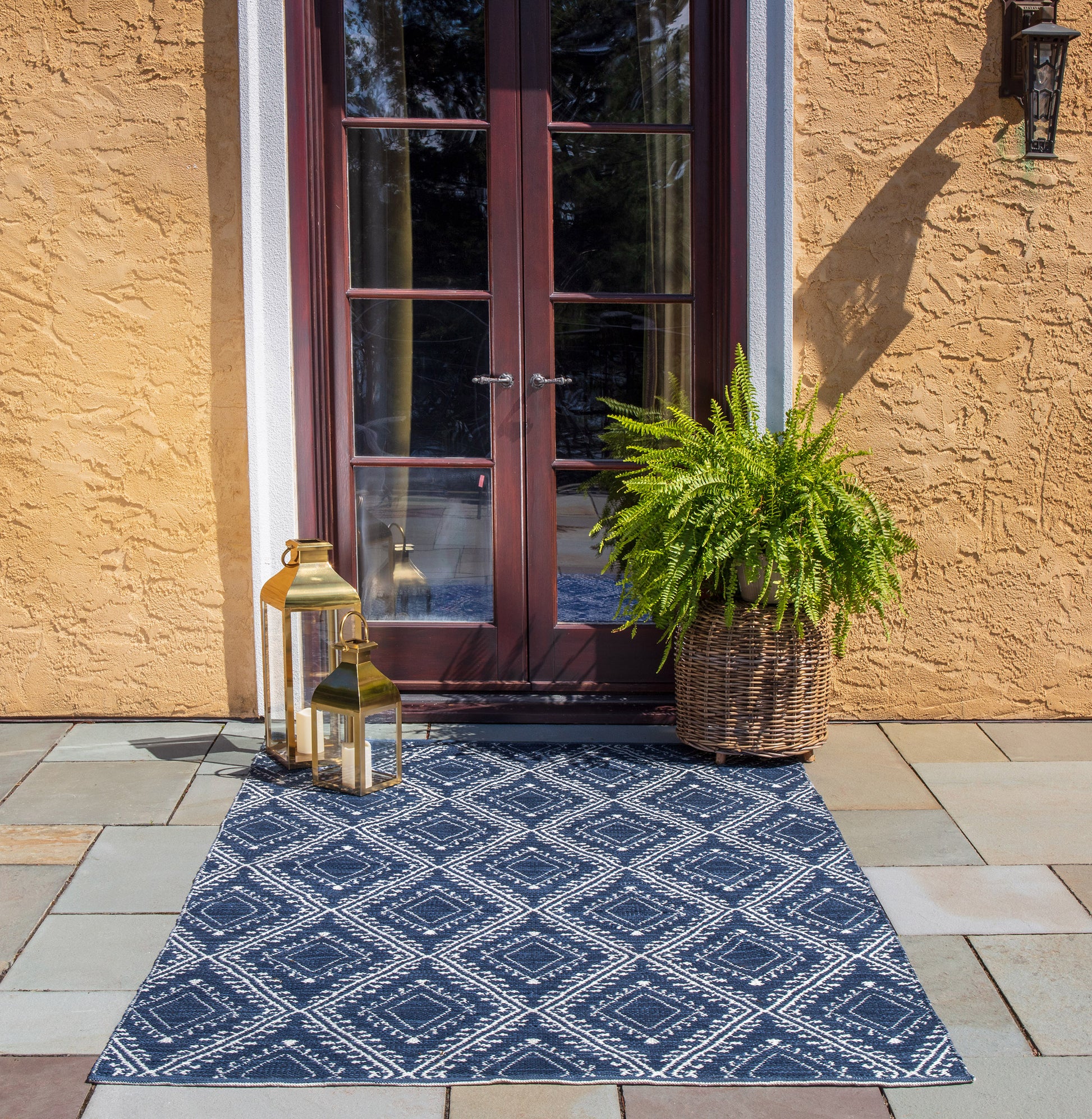 Navy outdoor rug with white diamond pattern in front of brown-framed glass doors; brass lanterns and a wicker fern planter.