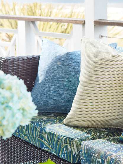 Outdoor wicker sofa with blue leaf-patterned cushions and blue and beige textured pillows on a sunlit porch.