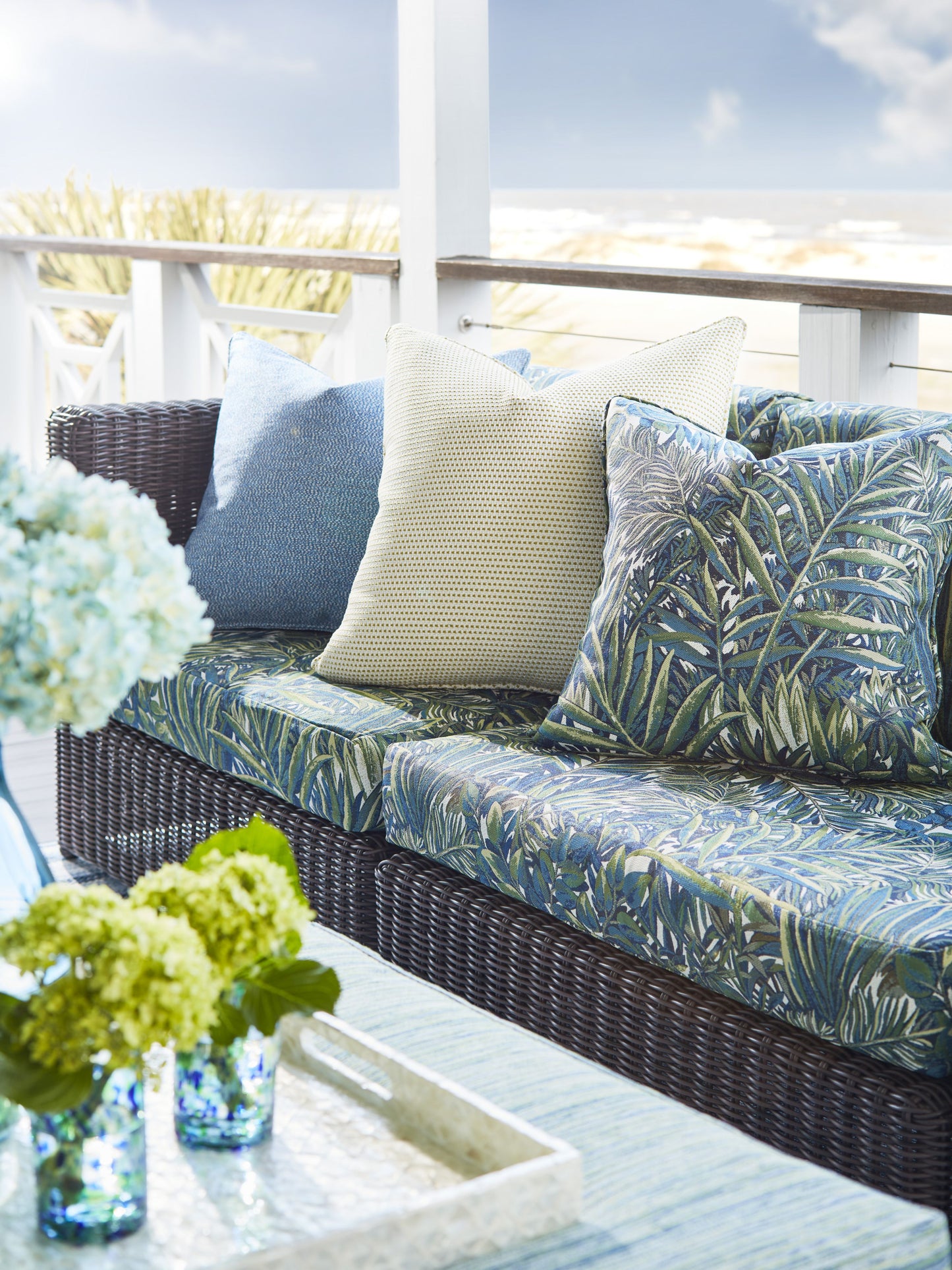 Wicker outdoor sofa with tropical leaf cushions, beige throw, blue pillow; glass coffee table with blue hydrangea vase.