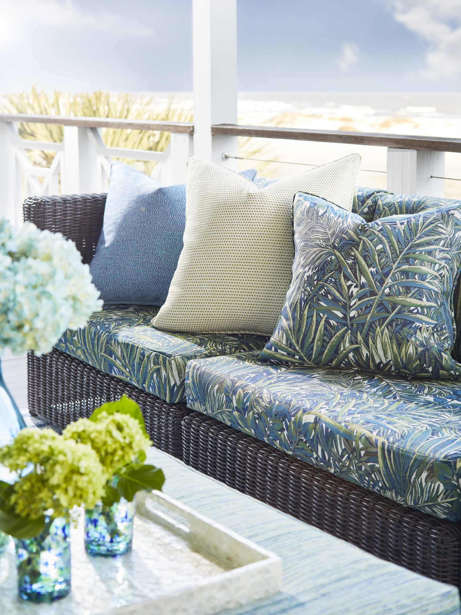 Wicker outdoor sofa with tropical leaf cushions, beige throw, blue pillow; glass coffee table with blue hydrangea vase.