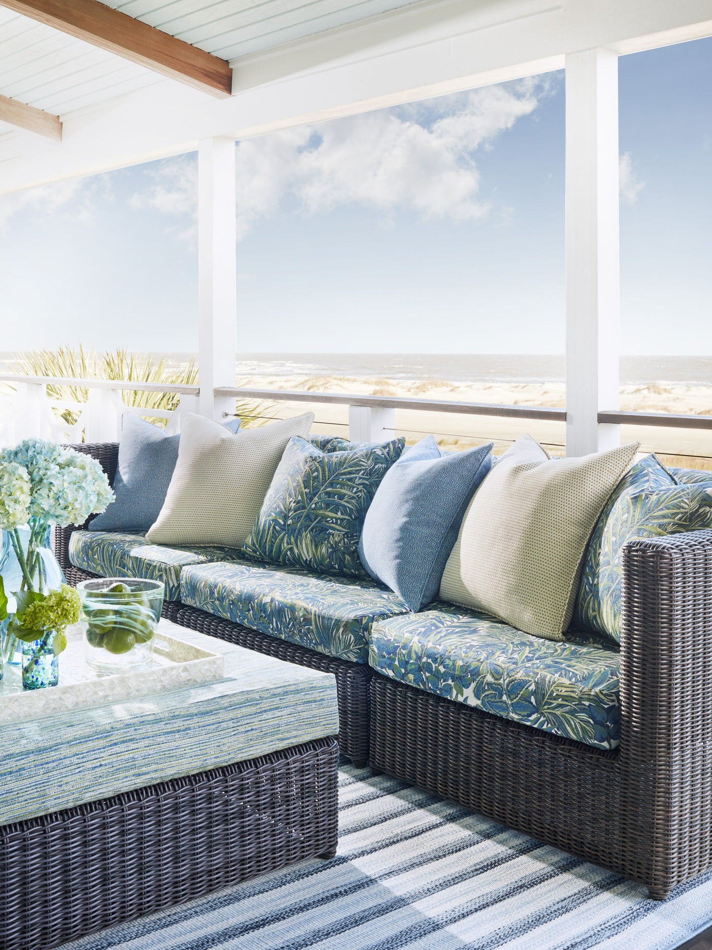 Coastal outdoor wicker sofa set with blue water cushions, glass-top coffee table, striped rug on a shaded patio.