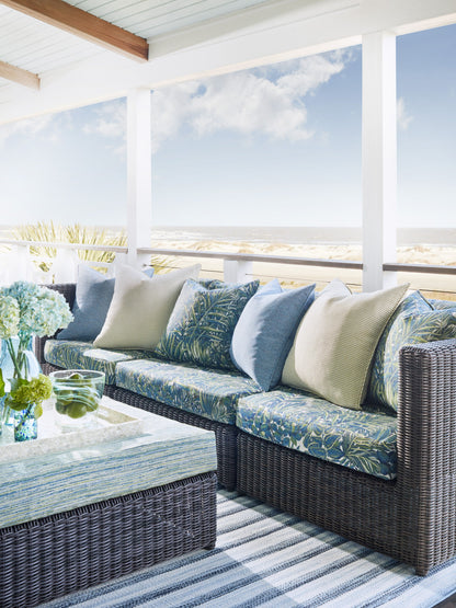 Coastal outdoor wicker sofa set with blue water cushions, glass-top coffee table, striped rug on a shaded patio.