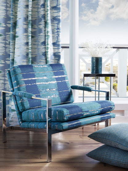 Blue patterned lounge chair with chrome frame, teal cushions, beside a side table with a blue vase in a sunlit room.