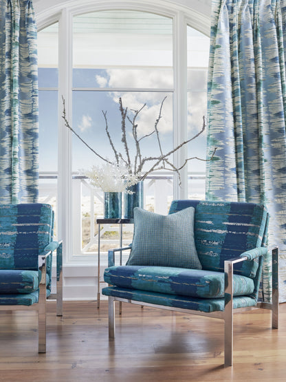 Blue upholstered lounge chairs with metal frames, light blue pillow, side table with white blossoms by arched window.