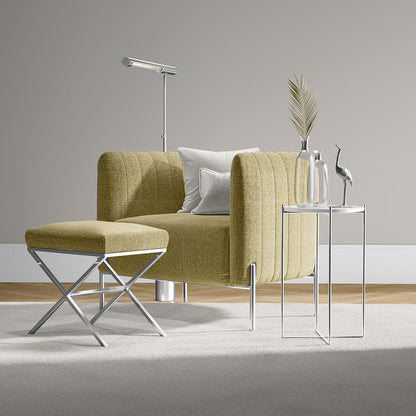 Mustard-yellow upholstered armchair with matching ottoman and chrome legs, white cushions, beside a glass-top side table with vases and a tall floor lamp.
