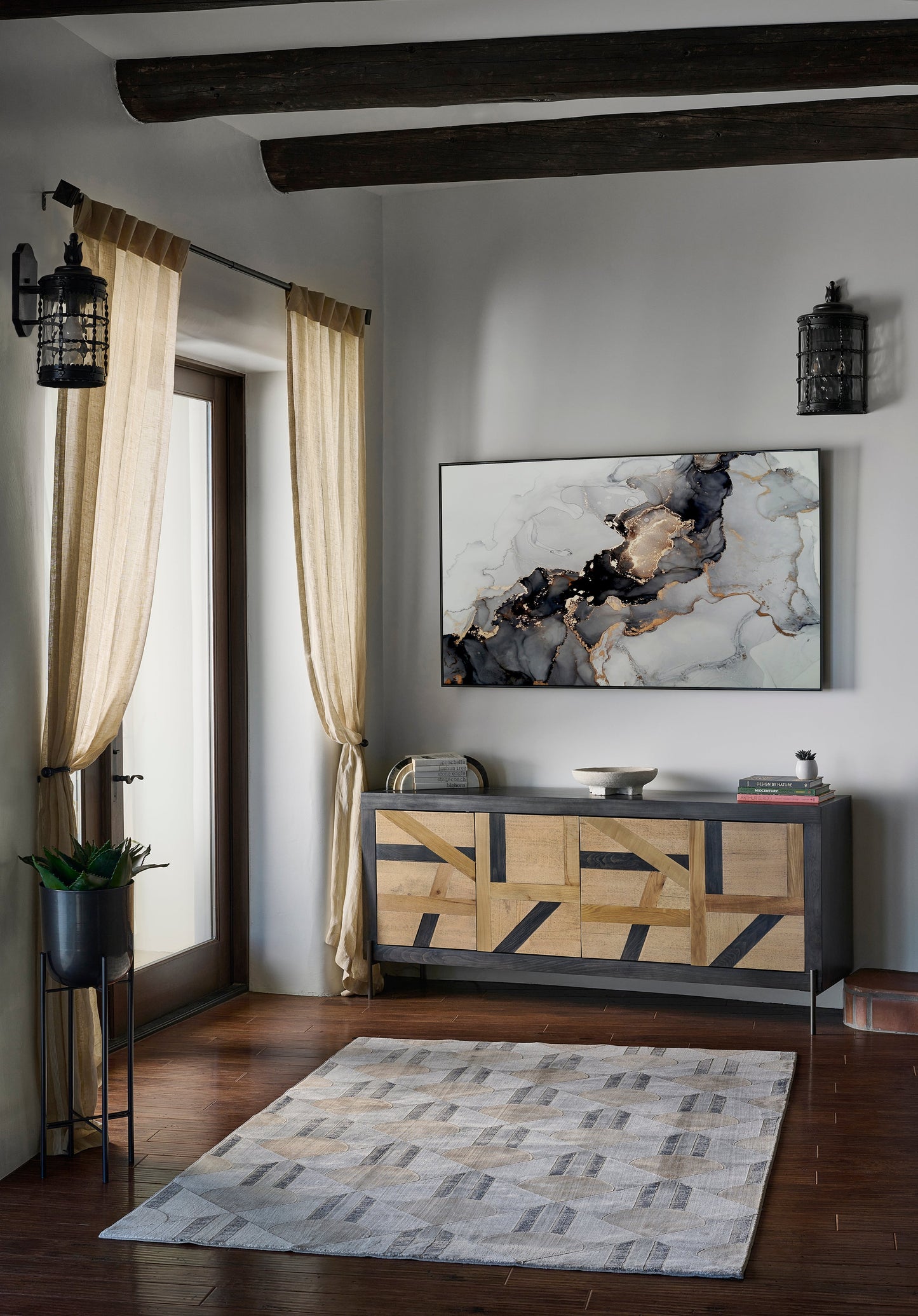 Living room corner with dark wood beams, beige curtains, a geometric wood cabinet, marble art, lantern sconces, plant, and rug.