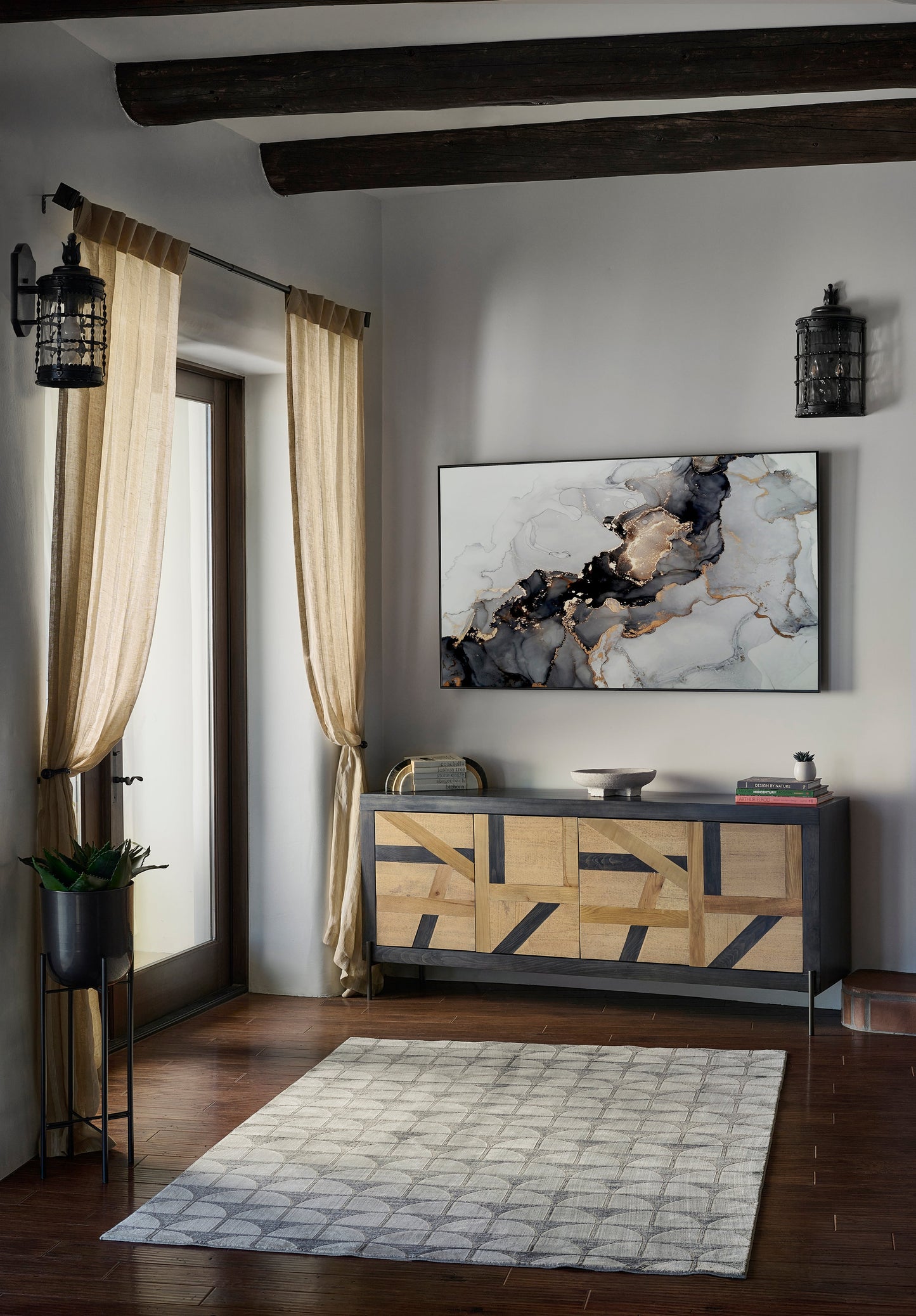 Cozy living room corner featuring a gray patterned rug, beige drapes, dark ceiling beams, and abstract art above a wood sideboard.