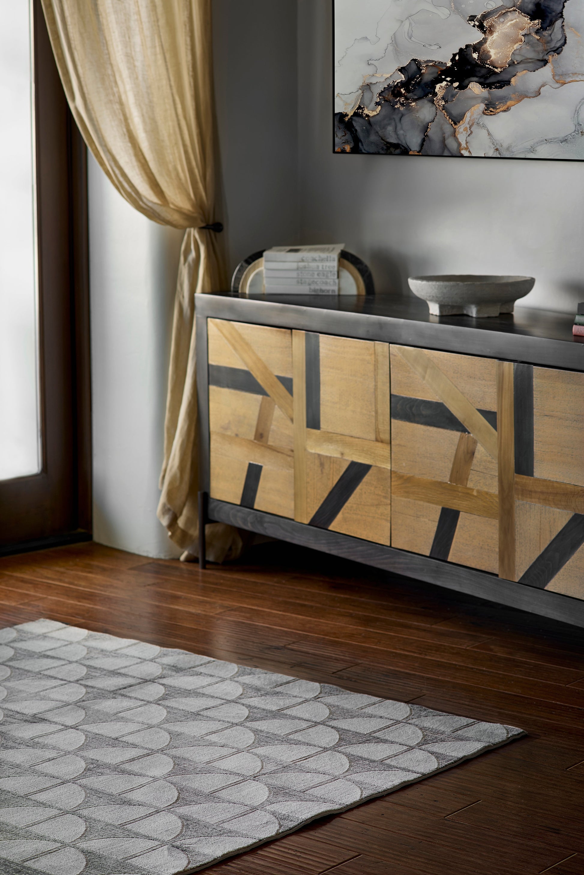Geometric wood sideboard with inlay and dark frame, gray top; décor bowls, wall art, gold curtain, and patterned gray rug.