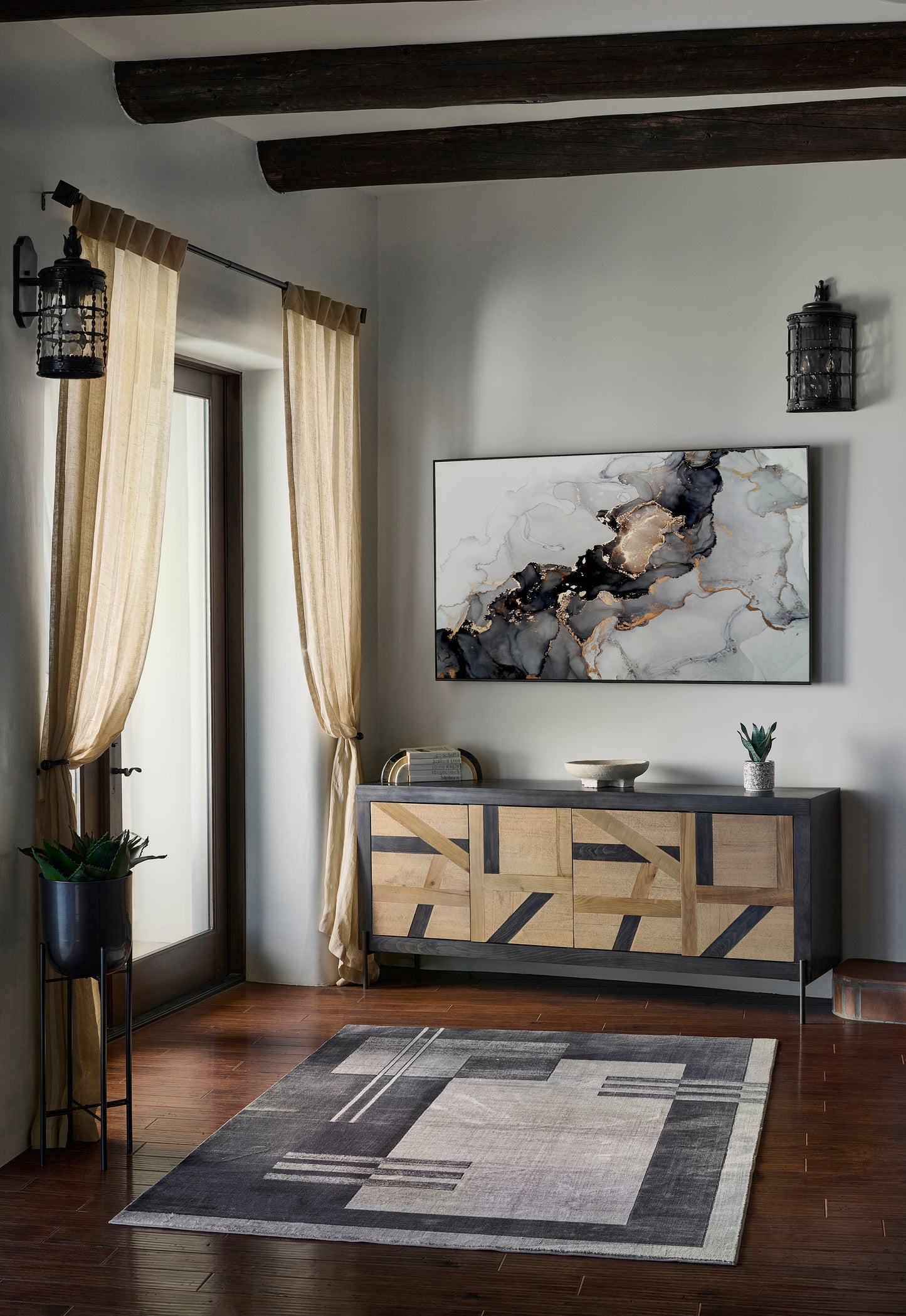 Rustic living room with geometric wood sideboard, abstract art, sheer beige curtains, black lanterns, and a gray rug.