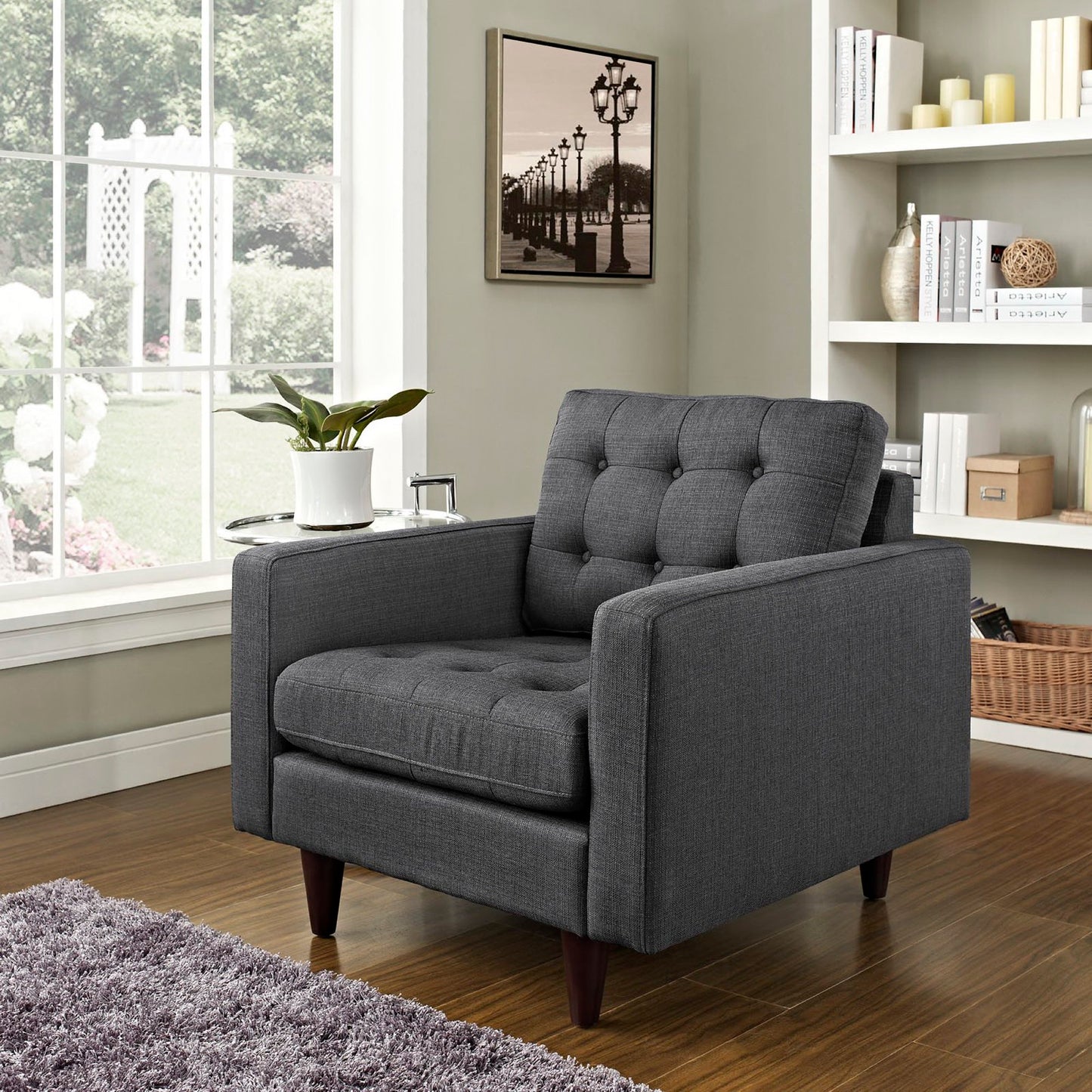 Dark gray tufted fabric armchair with square arms and tapered wooden legs in a sunlit living room with a bookshelf.