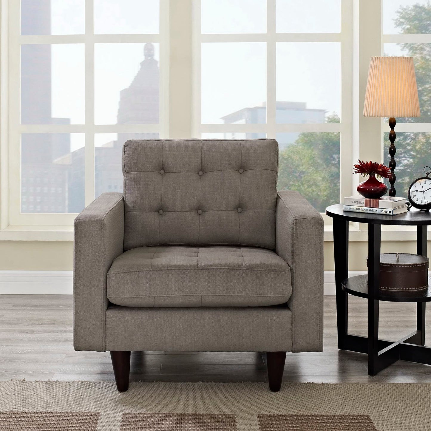 Grey tufted fabric armchair with square arms and dark wooden legs in a bright living room beside a round side table.