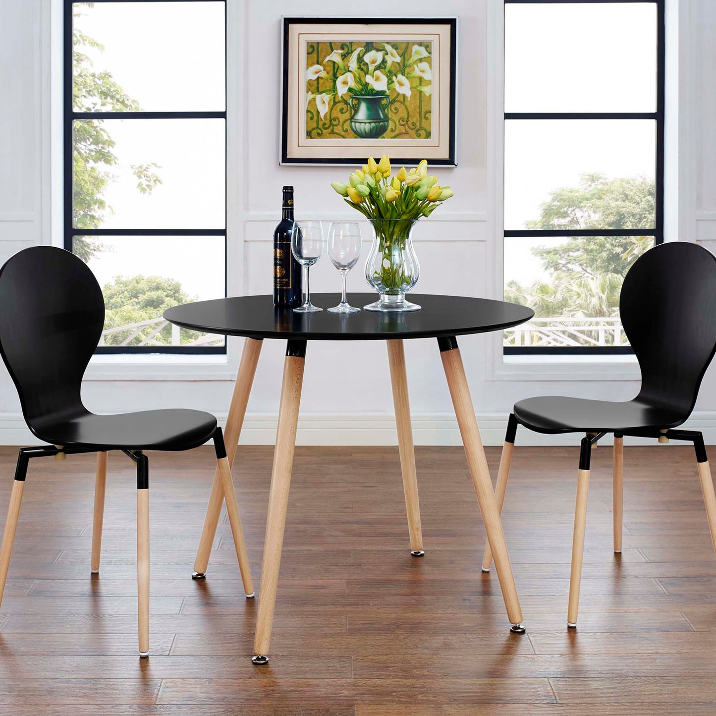 Round black dining table with natural wood legs, two black molded chairs; wine bottle, two glasses, and a vase of yellow tulips.