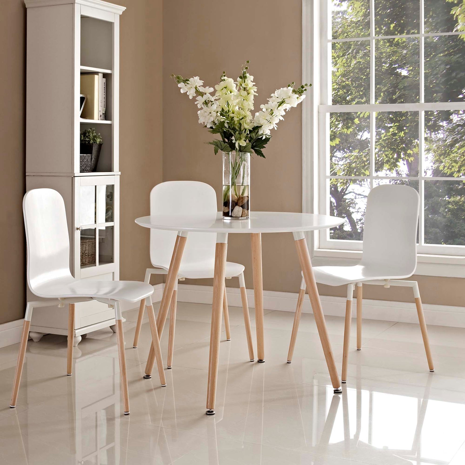 White round dining table with light wood legs, three white chairs, a vase of white flowers, in a sunlit dining room.
