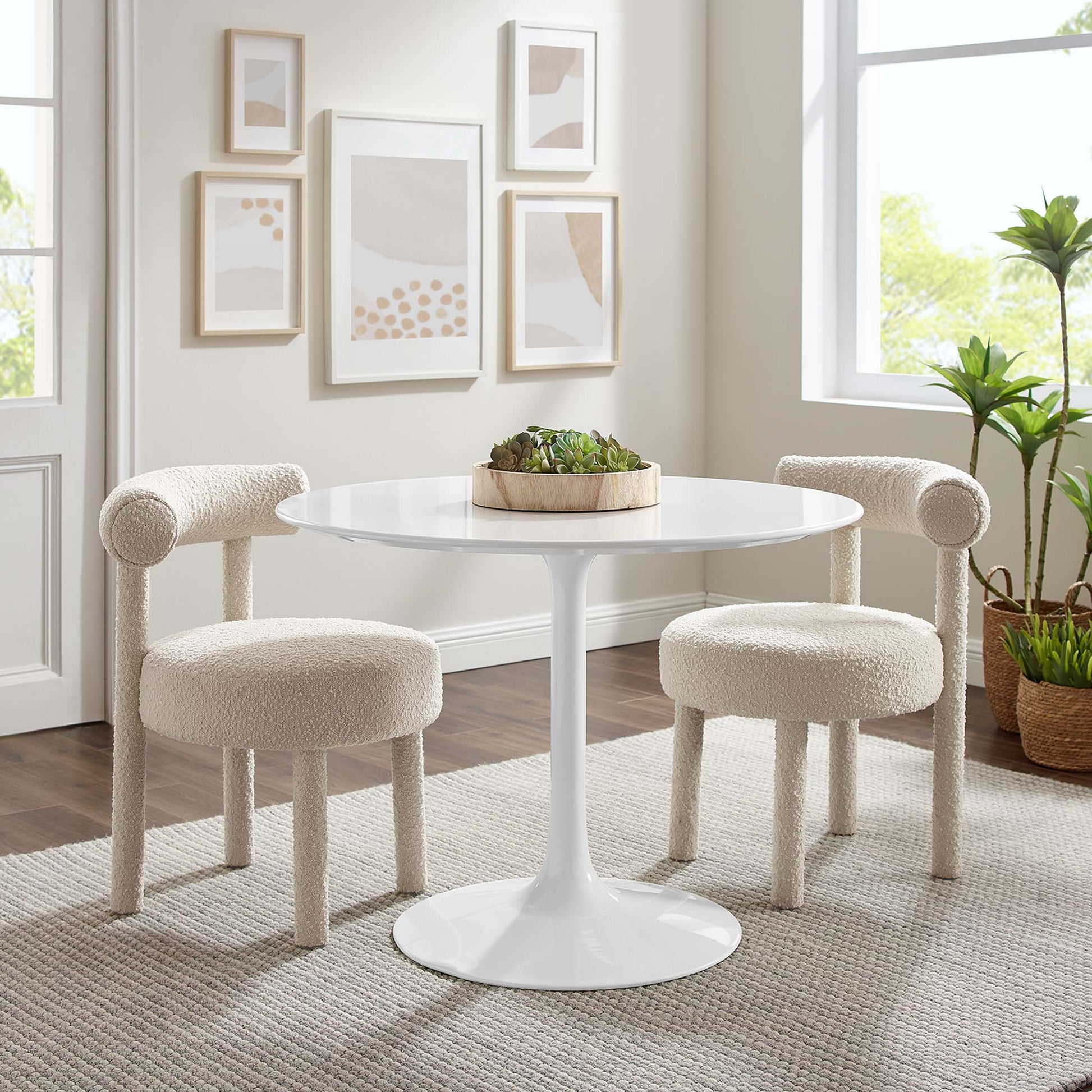 White round pedestal table with tulip base, two cream boucle armchairs, beige rug, wall art, and houseplants.