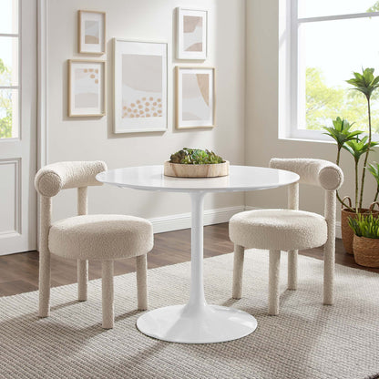 White round pedestal table with tulip base, two cream boucle armchairs, beige rug, wall art, and houseplants.