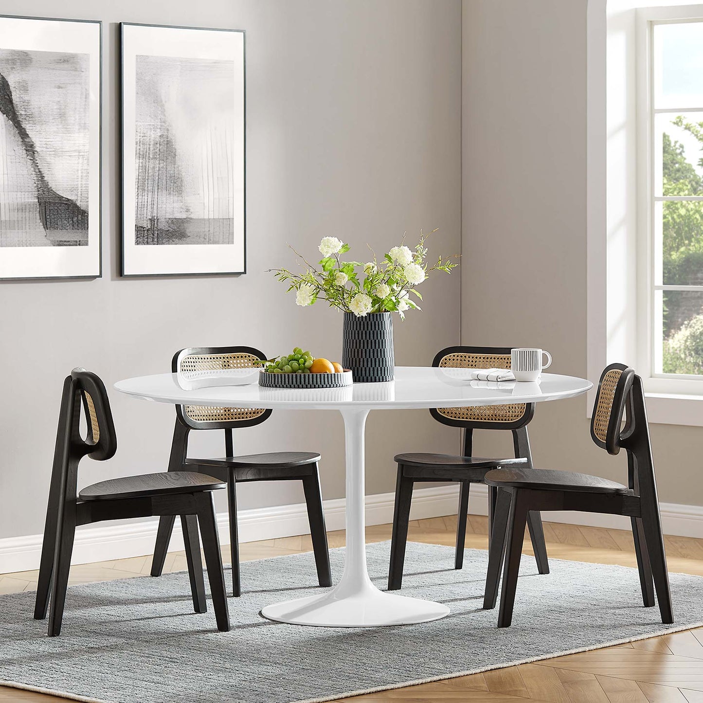 Round white tulip table with four dark wood chairs featuring cane backs; vase of white flowers, gray rug, modern dining set.
