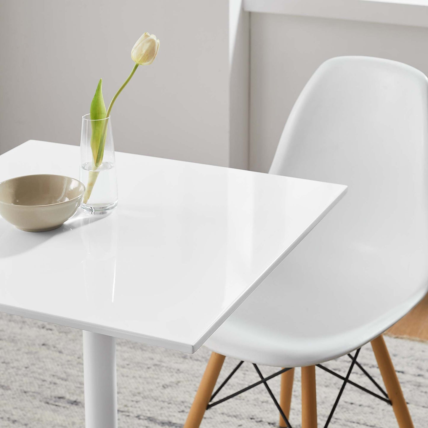 White glossy square dining table with a glass vase holding a white tulip and beige bowl; molded chair with wooden legs.