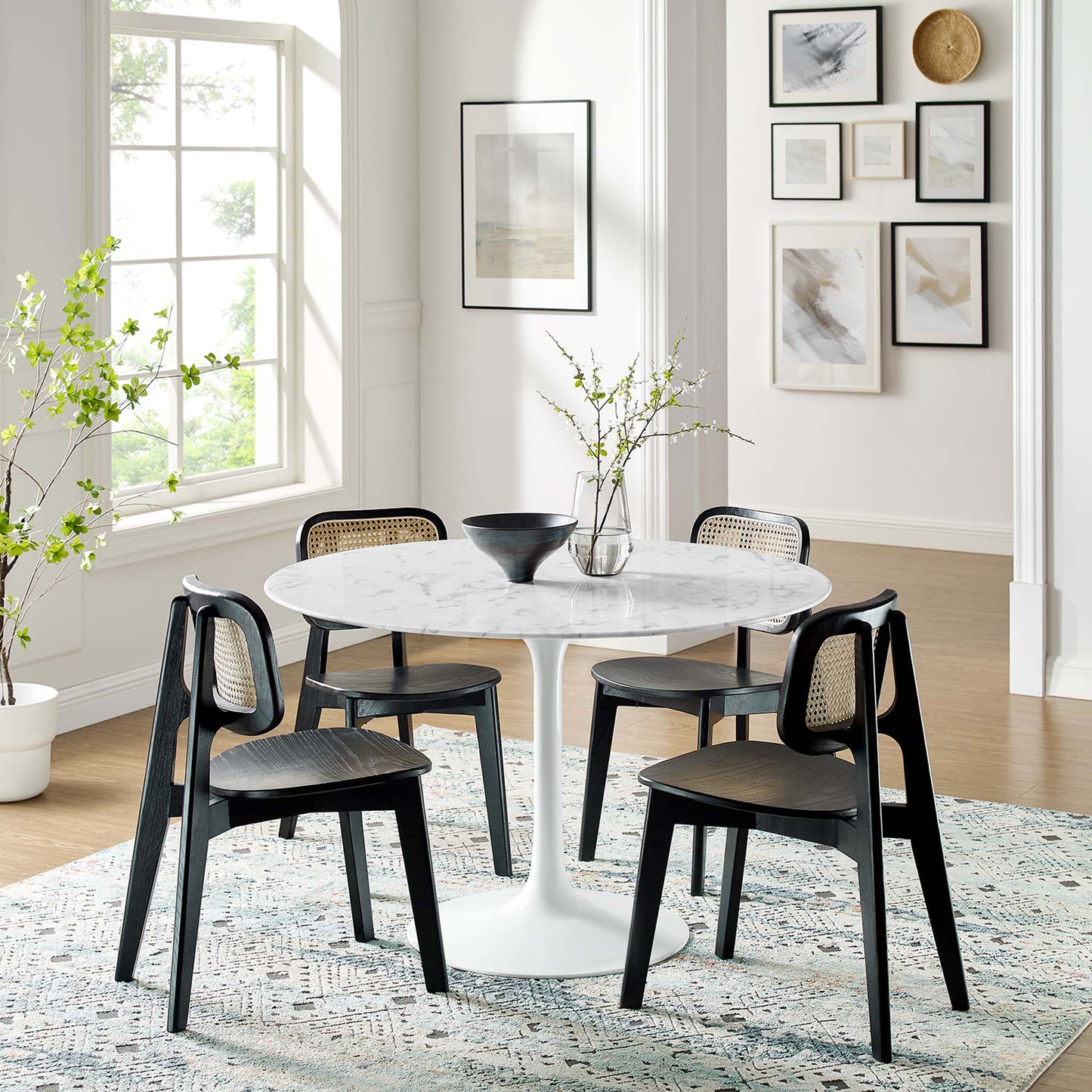 Round white marble dining table with pedestal base and black cane-back chairs, vase with branches in a bright modern dining room.