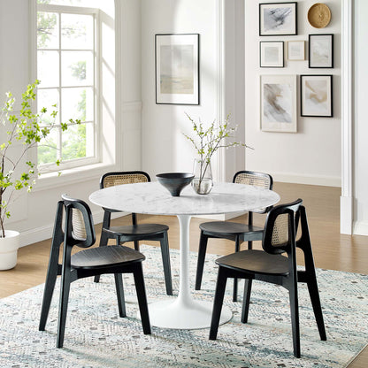 Round white marble dining table with pedestal base and black cane-back chairs, vase with branches in a bright modern dining room.