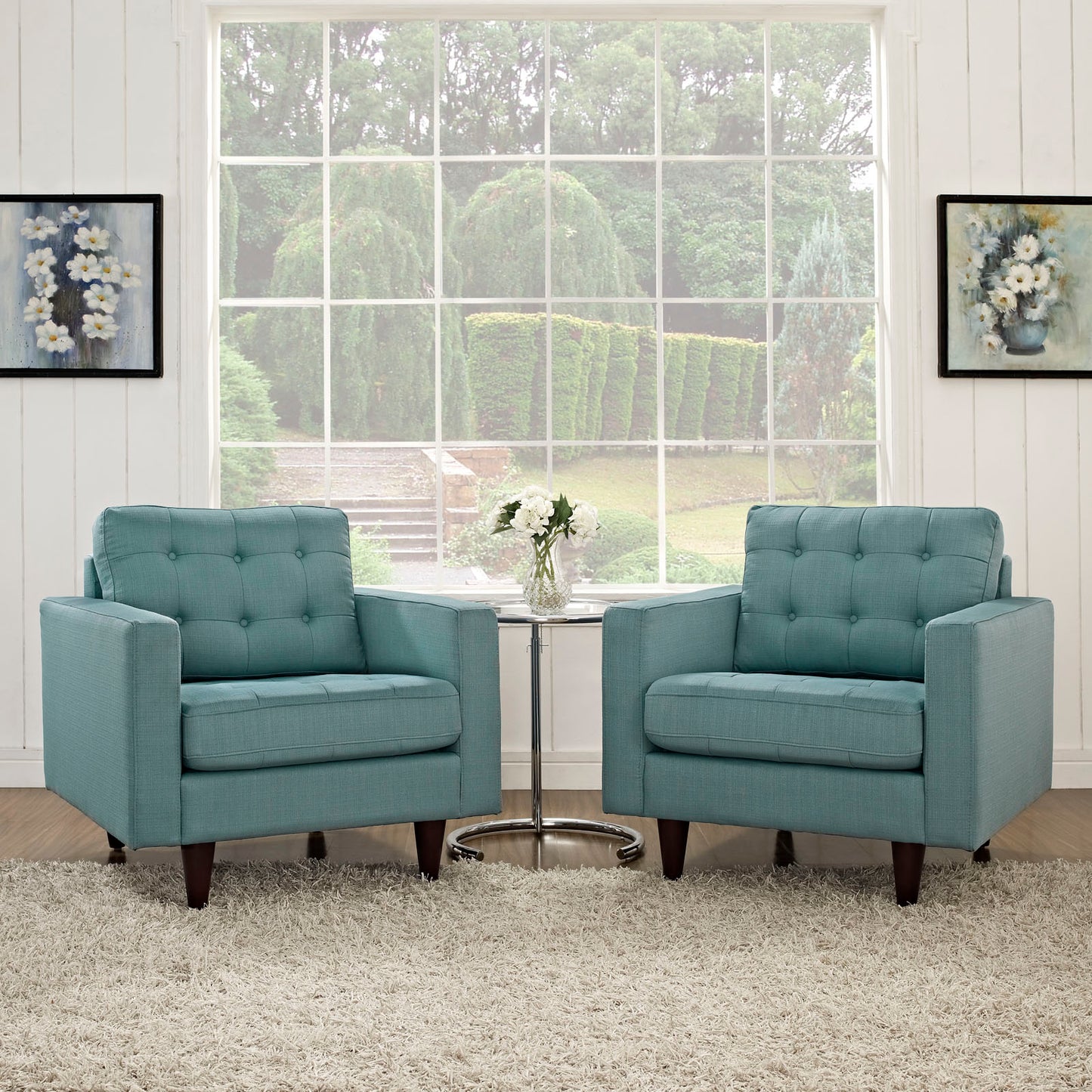 Two teal tufted armchairs with dark wooden legs flank a round glass coffee table in a bright, airy living room.