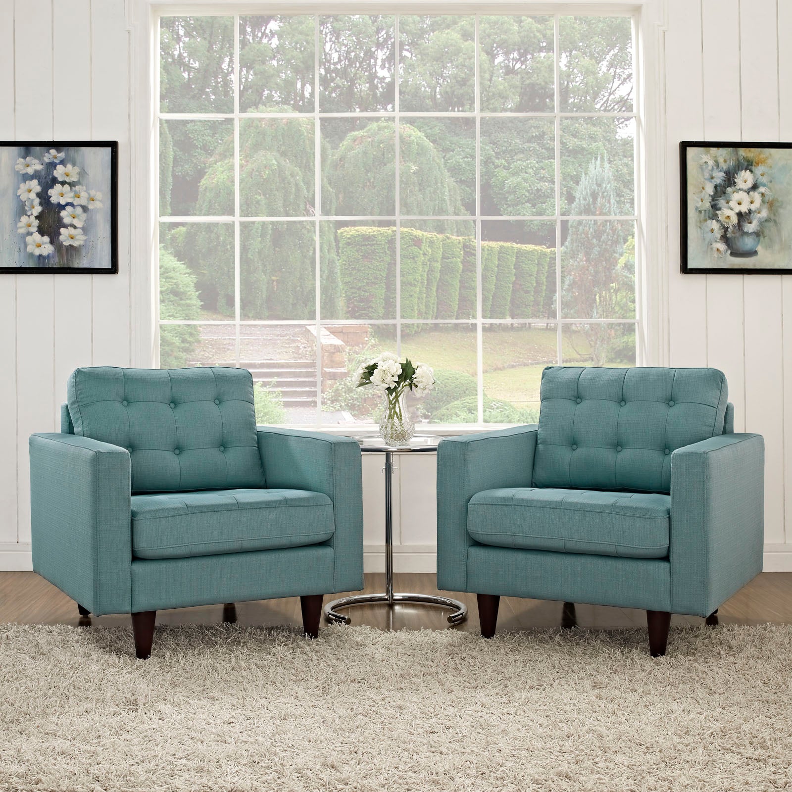 Two teal tufted armchairs with dark wooden legs flank a round glass coffee table in a bright, airy living room.