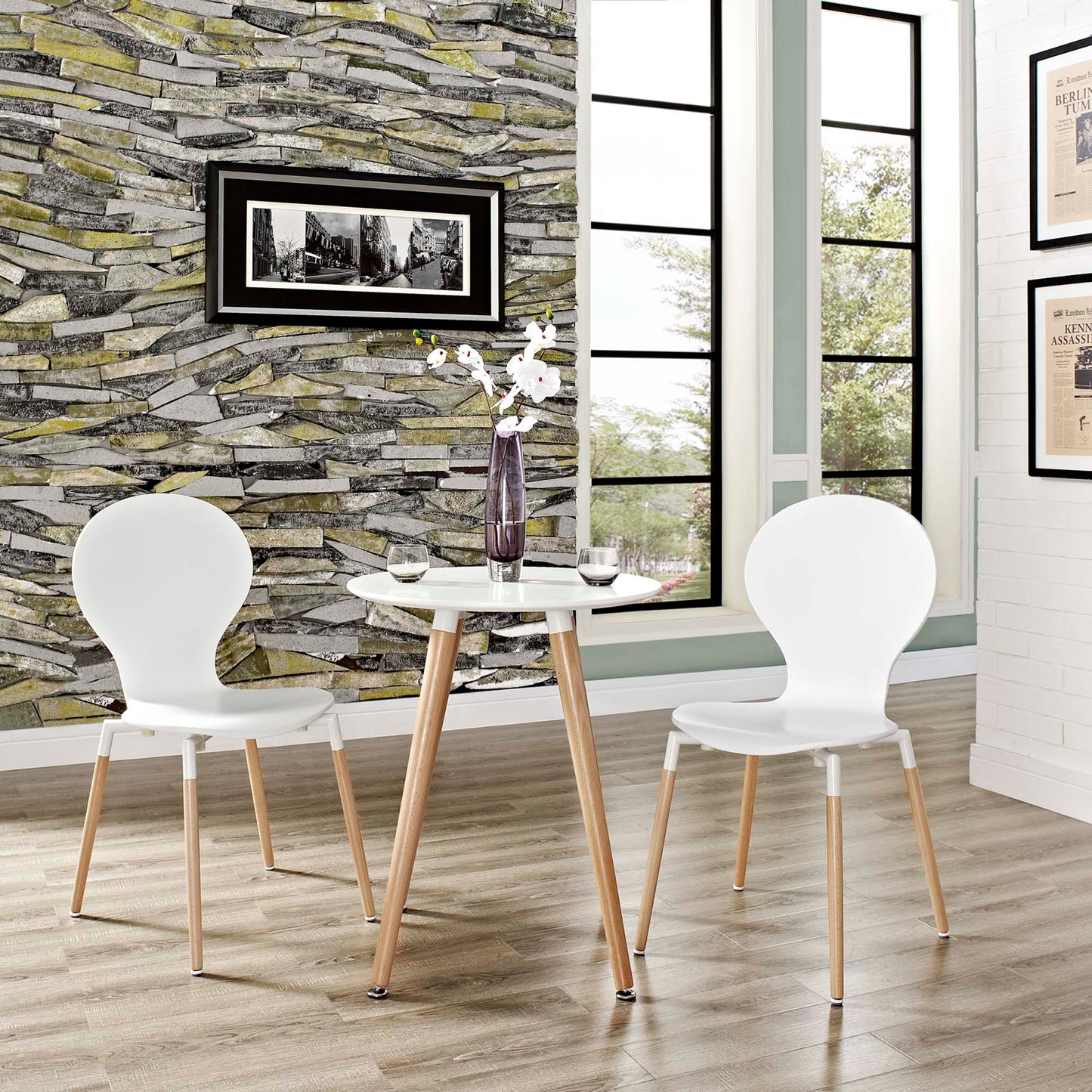 White round table with light wood legs, two white modern chairs, vase of flowers, stone accent wall and large window.