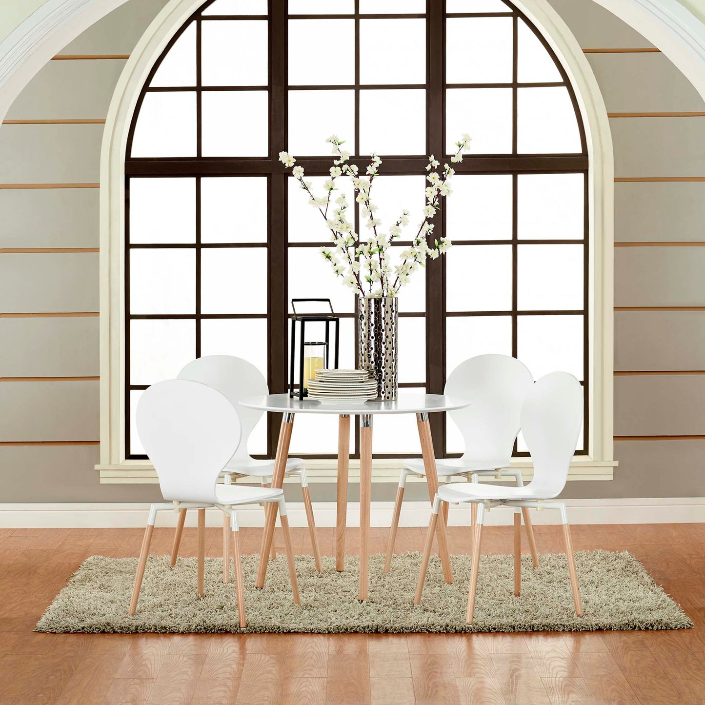 White round dining table with light wood legs and six white molded chairs, vase centerpiece on a rug beside an arched window.