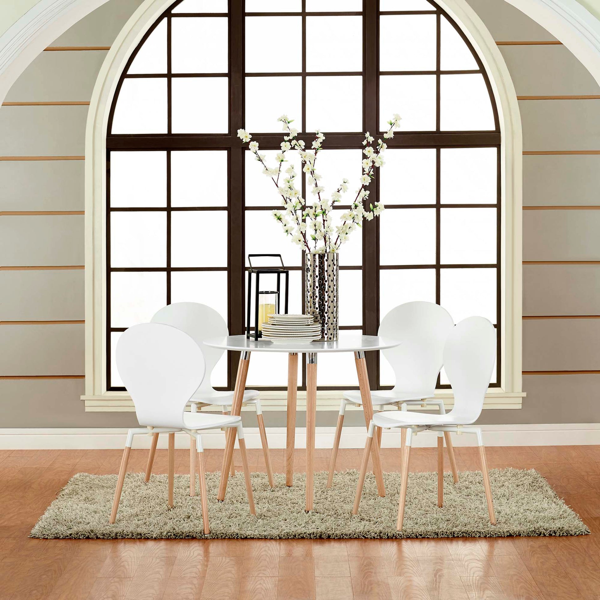 White round dining table with light wood legs and six white molded chairs, vase centerpiece on a rug beside an arched window.