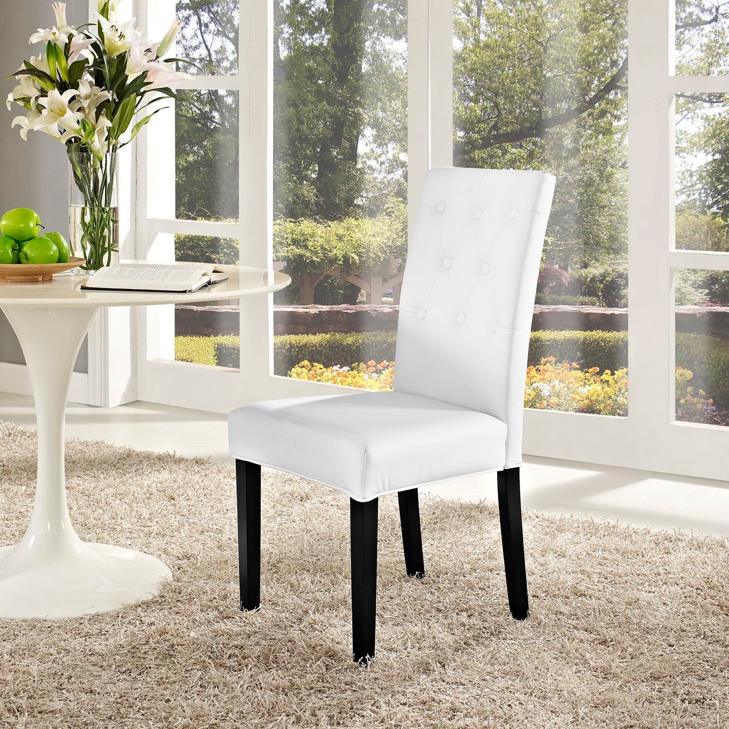 White tufted dining chair with black legs in a bright sunlit room beside a round white table with flowers and apples.