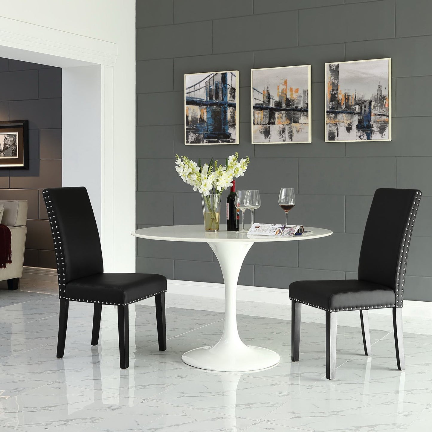 White round pedestal table with two black nailhead-trim chairs, vase of white flowers, wine glasses.