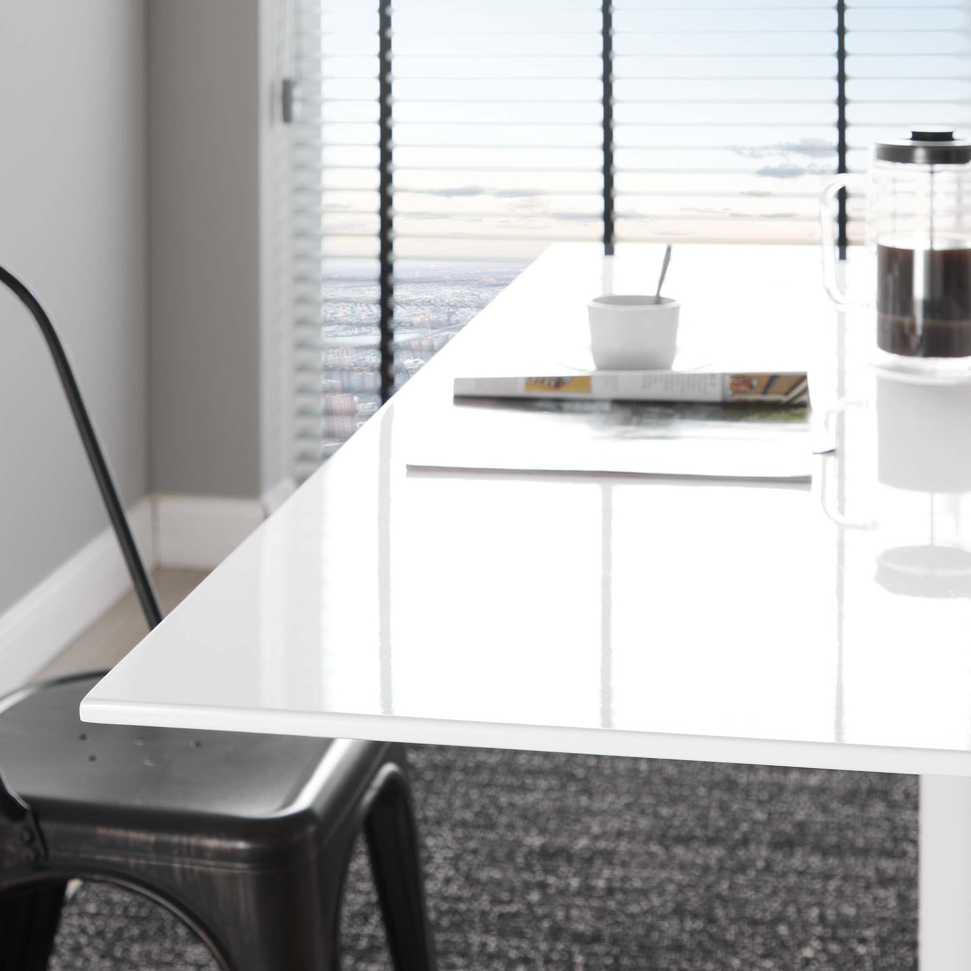 Glossy white rectangular table with chrome legs in a bright modern room; mug, magazine, spoon & French press on top.