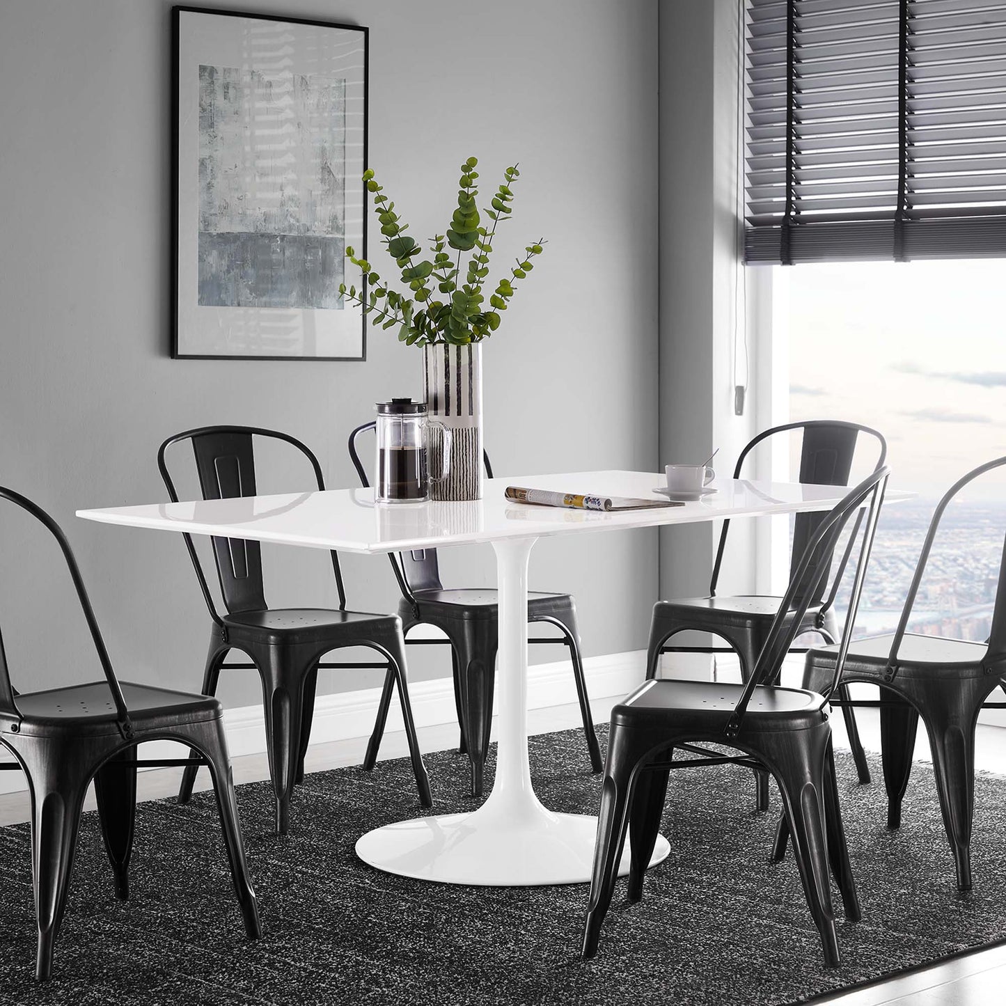 White round pedestal table with black metal chairs in a grey dining area; vase of greenery, magazine and cup by the window.