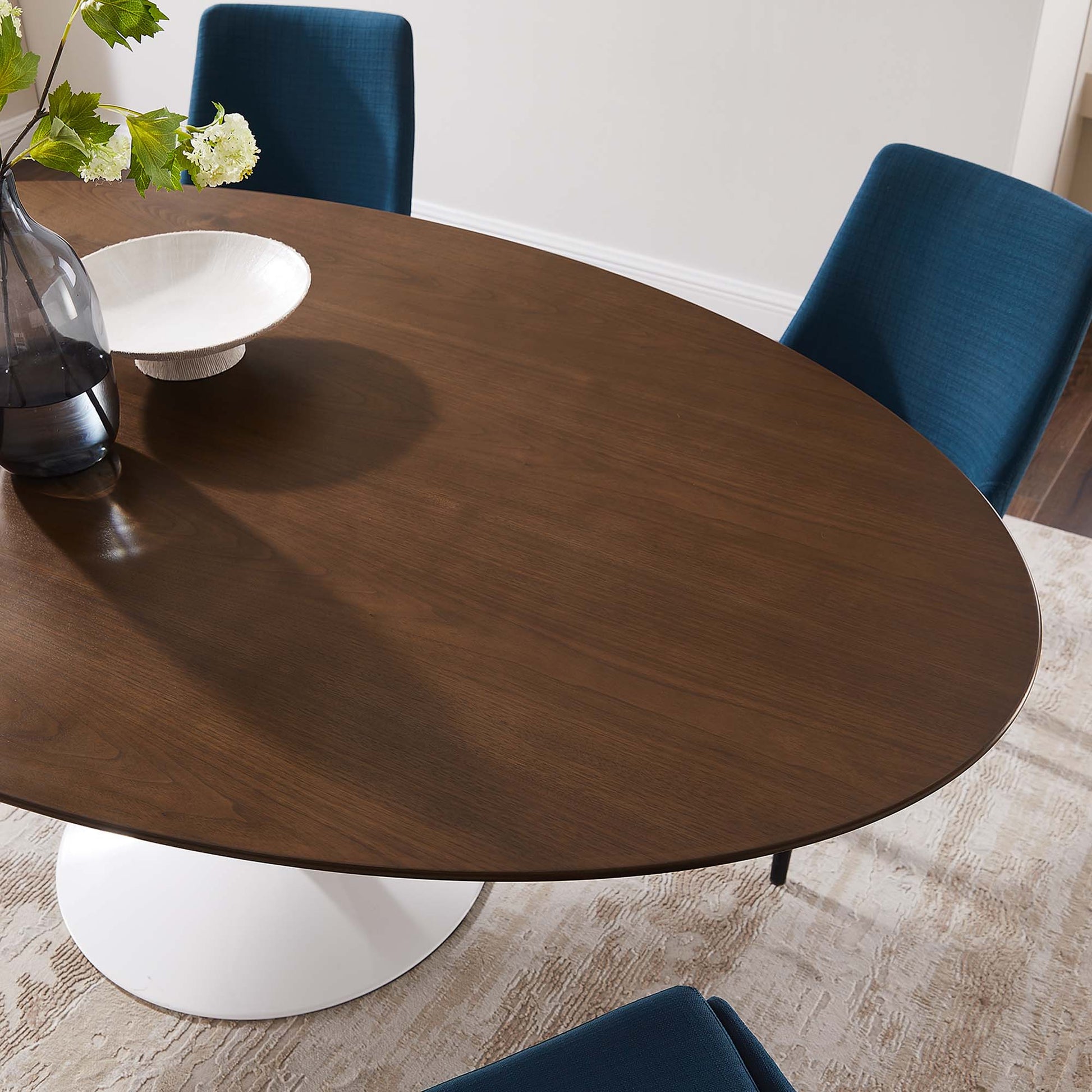 Round walnut wood dining table with white pedestal base, blue upholstered chairs, vase with greenery and white bowl.