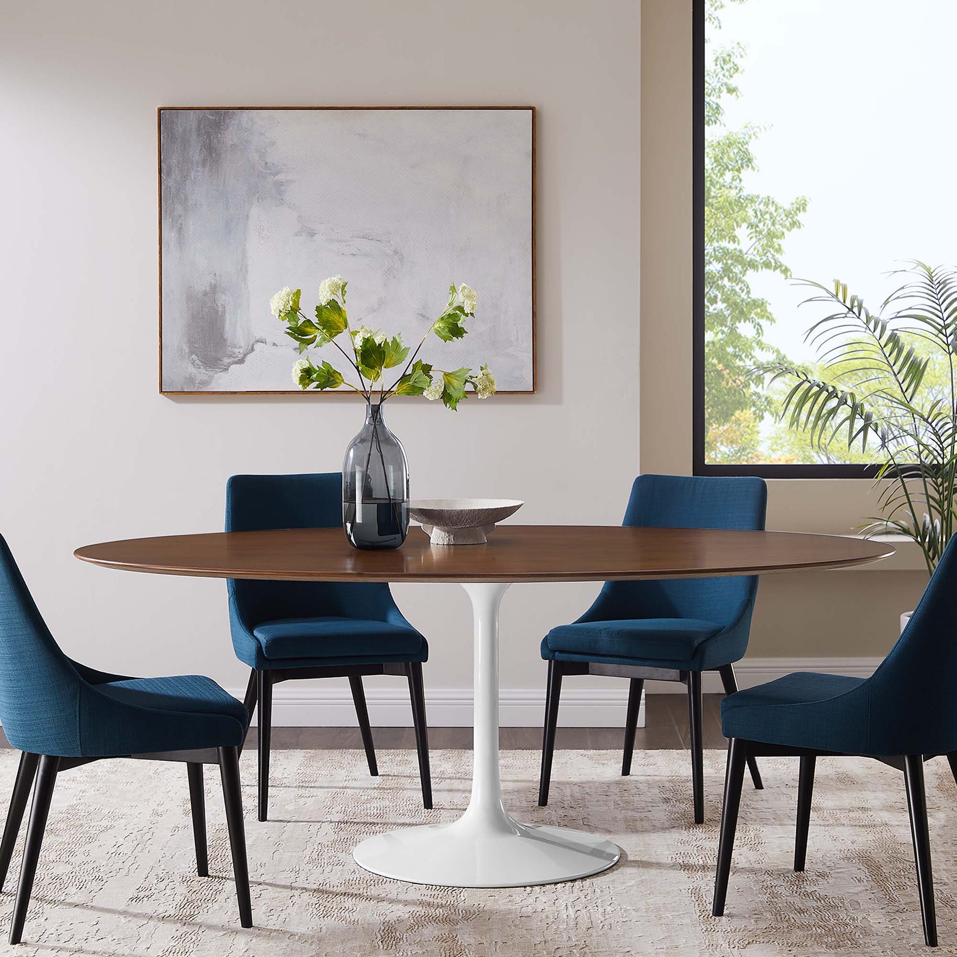Round wooden dining table with white pedestal base, blue upholstered chairs, glass vase with green stems, abstract wall art