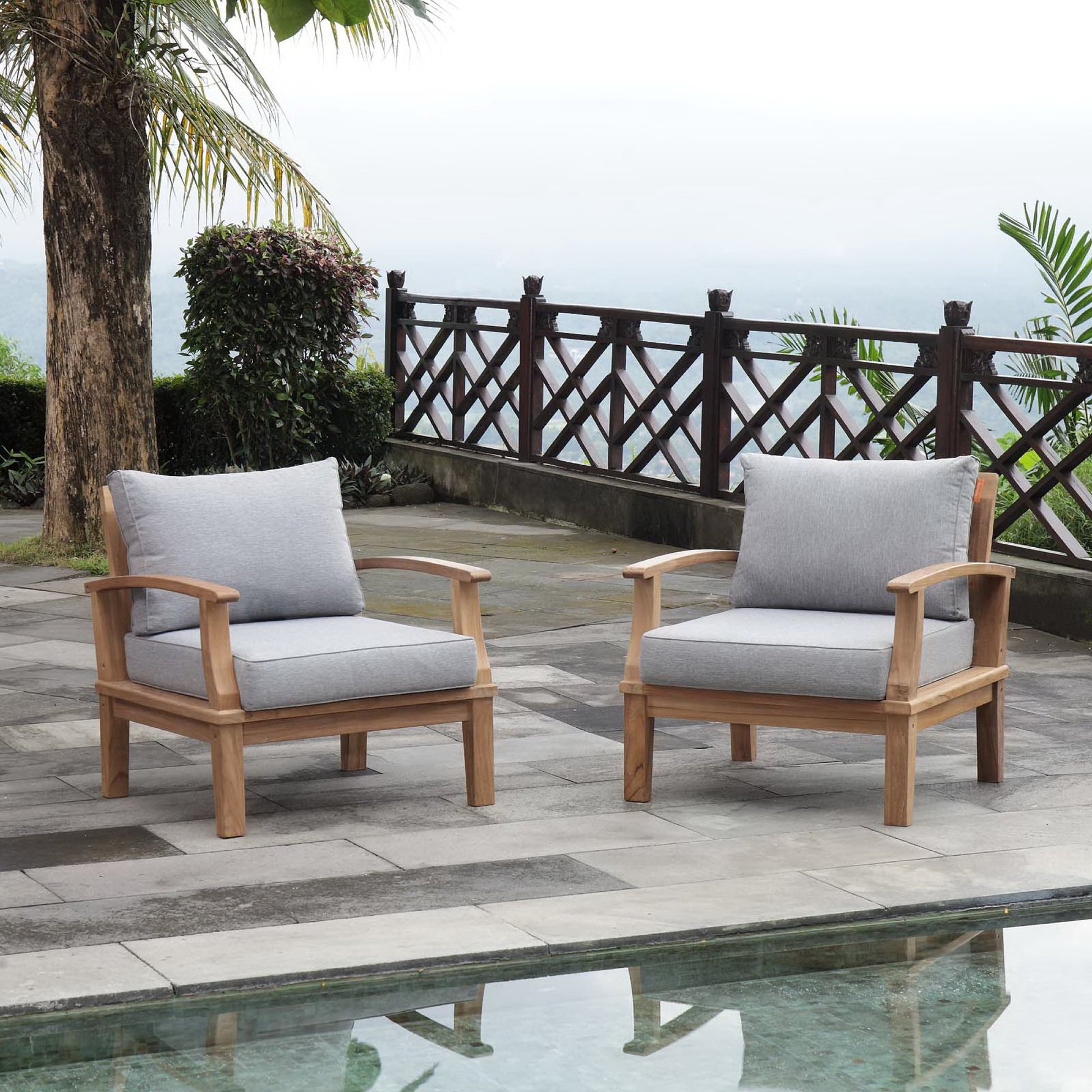 Natural wood outdoor armchairs with light gray cushions on a tiled patio beside a pool and wooden lattice railing.