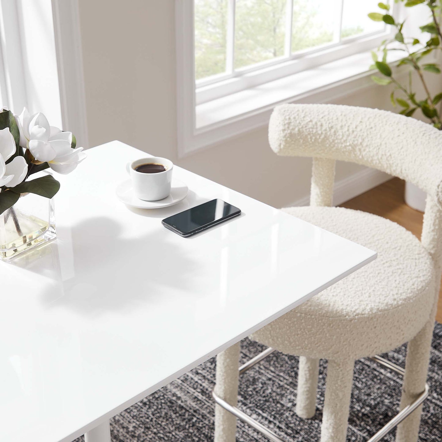 White glossy table with coffee on saucer, smartphone, vase of white flowers, cream textured chair, bright window.
